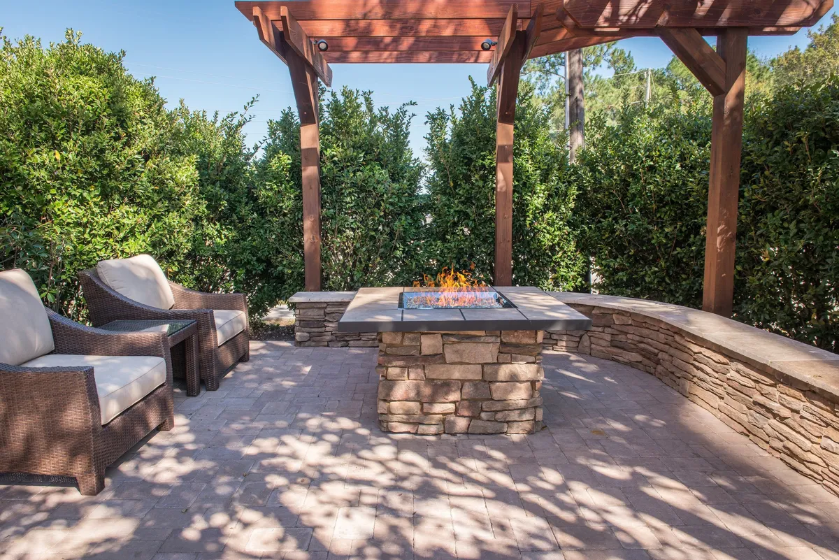 A cozy outdoor seating area centered around a fire pit, under a pergola structure. The space is designed for relaxing and socializing, perfect for unwinding after a workday. The area is surrounded by greenery, with comfortable chairs arranged for conversation or casual gatherings.