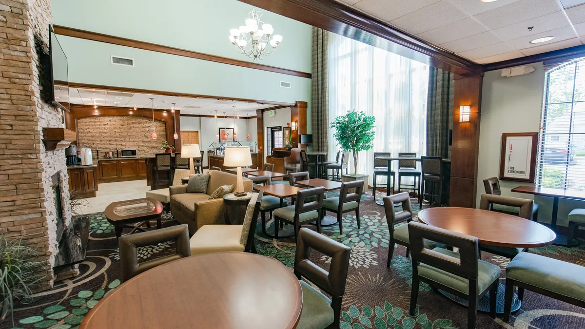 A spacious living area bathed in natural light from large windows. Plush, comfortable seating is arranged around a stone fireplace, with decorative plants adding warmth. The room also features a coffee station and multiple seating areas suitable for both relaxation and conversation. People can be seen working on laptops, reading, or enjoying coffee, creating a multi-functional and inviting atmosphere.
