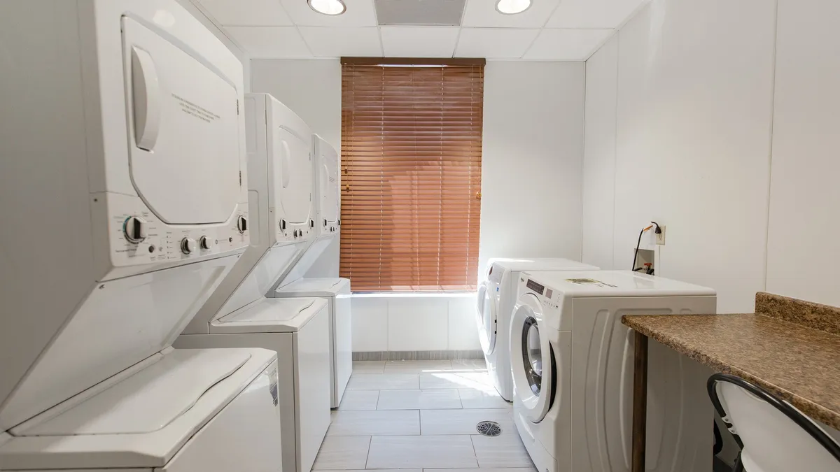 A tidy, functional laundry space with stacked washer and dryer units, a clean countertop, and sunlight filtering through window blinds. This practical area showcases storage and utility features essential for city living.
